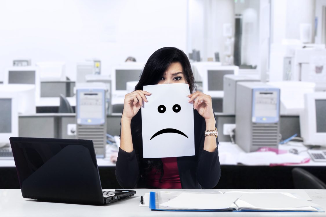 Annoying Coworker Habits Ranked Woman sits in office behind a sad face printed on a piece of paper