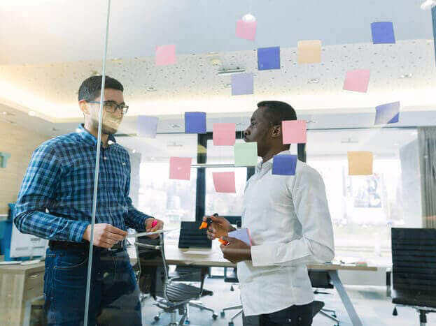 Two men talking in a modern startup office using a glass wall covered with sticky notes