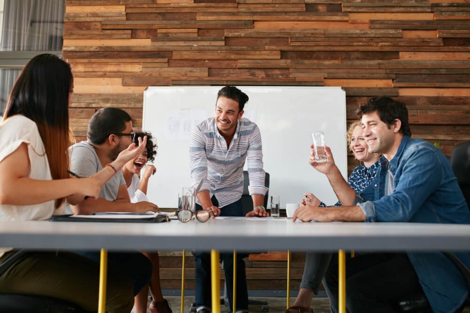 Team of men and women gathered around a table and whiteboard talking and business planning in a modern office space